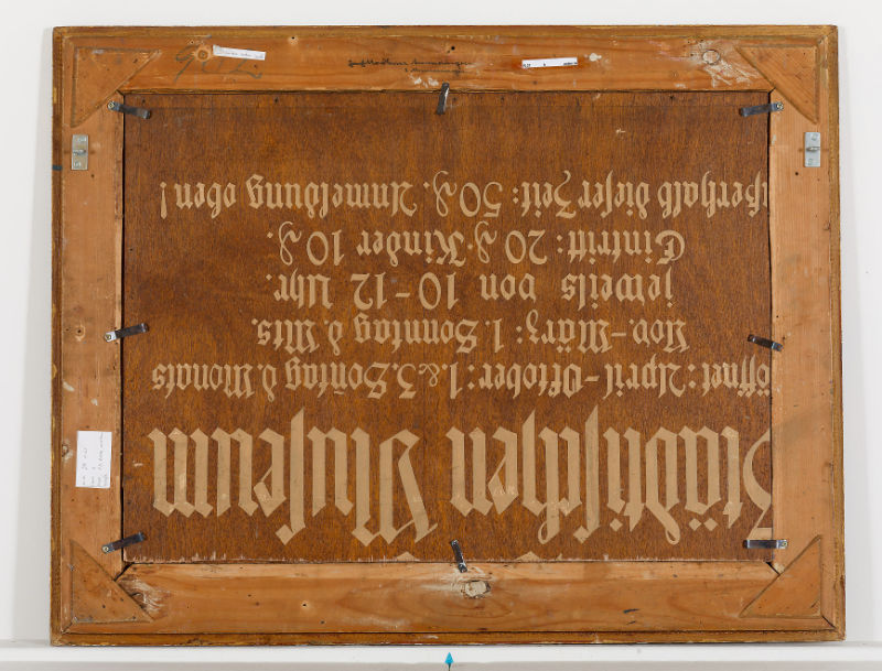 Die Rückseite eines gerahmten Bildes, das ursprünglich ein Schild für das Städtische Museum mit den Öffnungszeiten in gebrochener Schrift war
