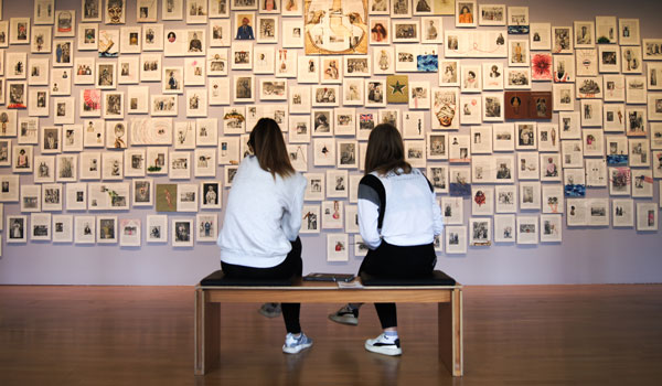 zwei junge Personen sitzen auf einer Bank vor einer Ausstellungswand mit vielen Bildern