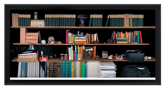 Fotografie eines Regals vor schwarzem Hintergrund mit Büchern, Dokumenten, technischen Geräten und Erinnerungsstücken