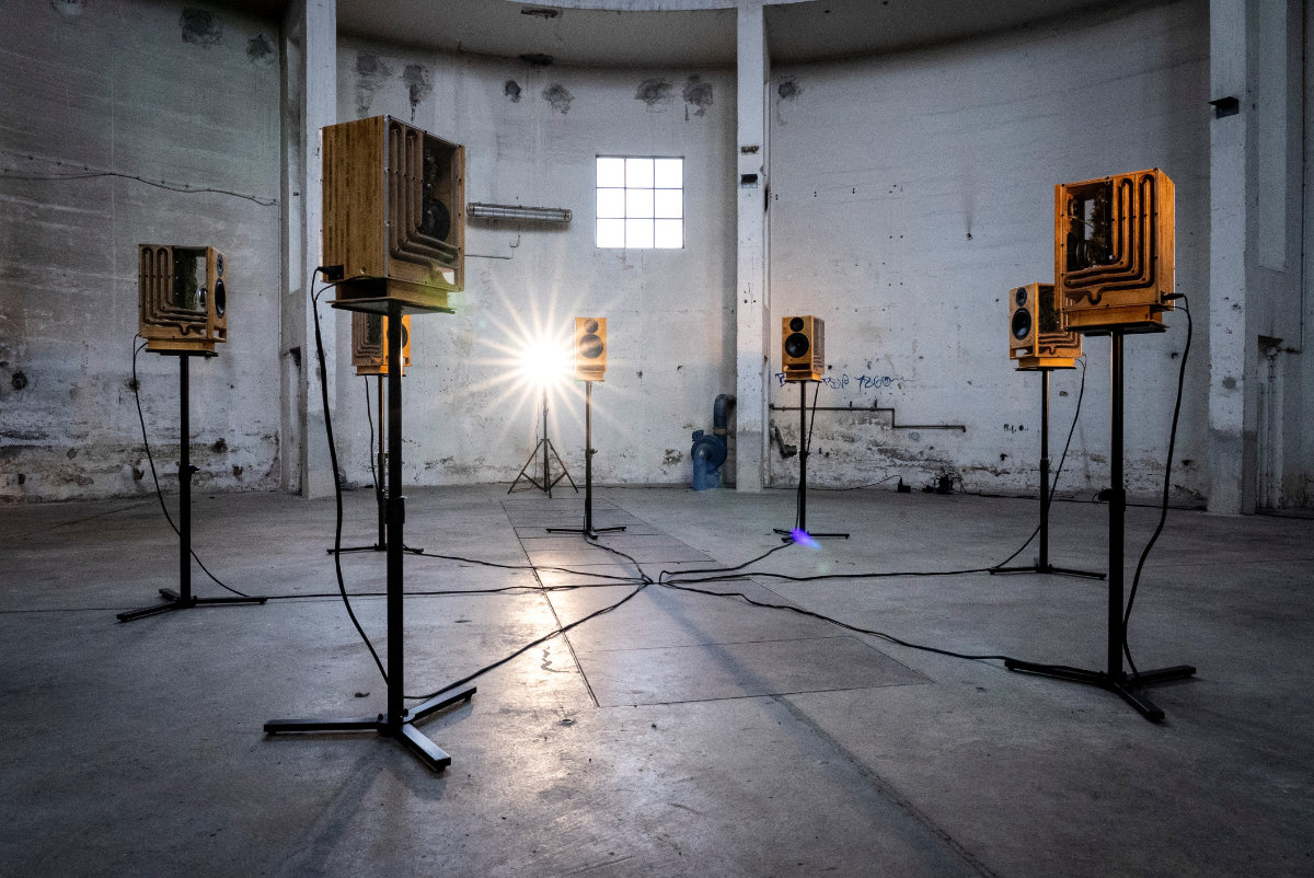 sieben im Kreis angeordnete Lautsprecherboxen auf Ständern in einem leeren, großen Raum, der wie eine Fabrikhalle wirkt