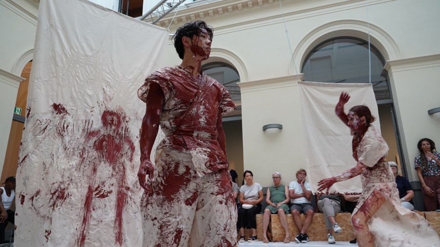 Tänzer bewegen sich zwischen weißen Tüchern. Die weiße Kleidund der Tänder und die Tücher sind mit roter Farbe verschmiert.