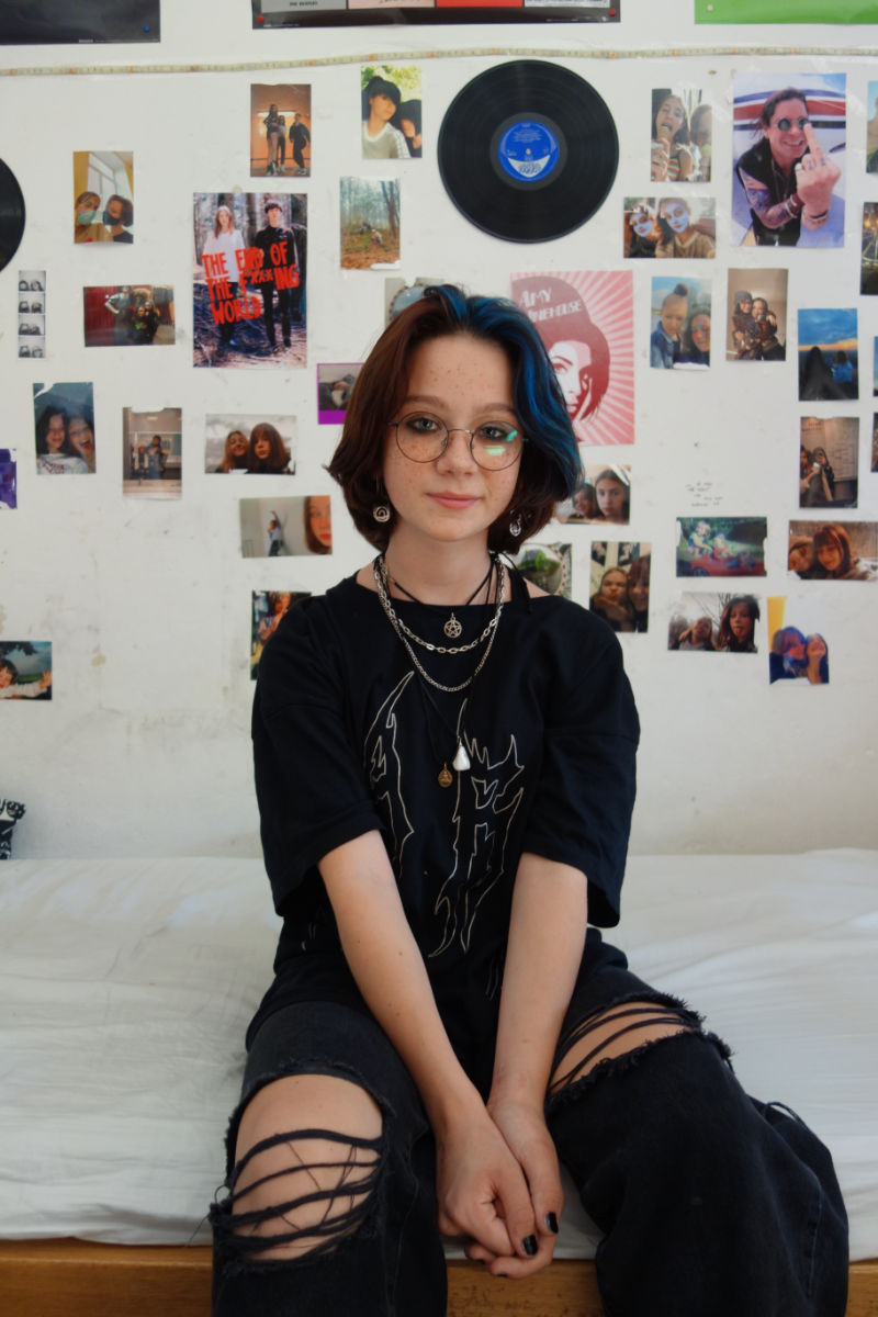 eine junge Frau in schwarzer Kleidung sitzt auf einem Bett vor einer Wand voller Fotos