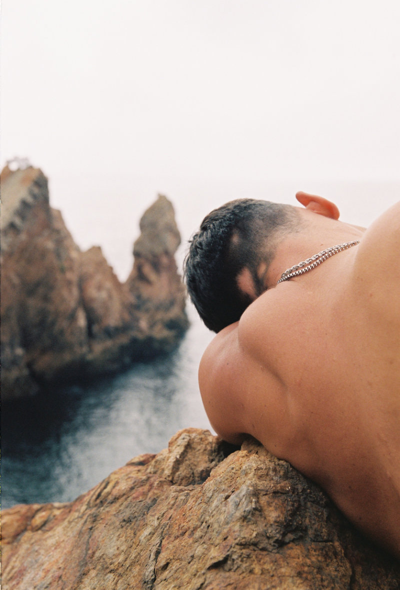 Die Ansicht des Rückens und Hinterkopfes eines jungen Mannes, der am Meer auf einem Felsen lehnt